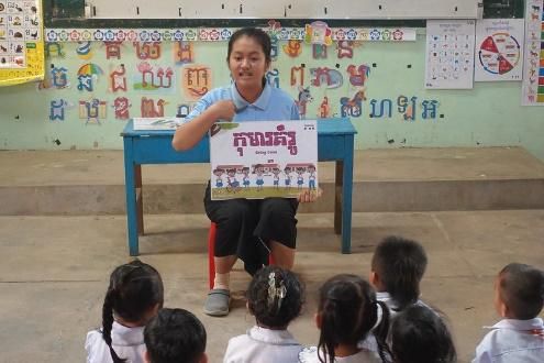 A teacher sits at a desk and holds up a picture card to children seated on the floor