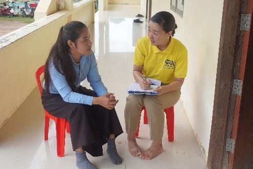 A staff member in yellow takes notes while talking with a teacher seated on a red plastic chair