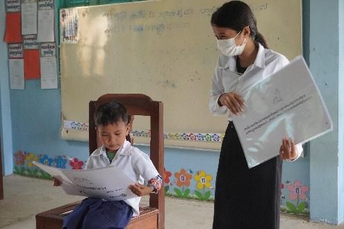 A teacher in a face mask holds up a large sheet beside a pupil reading a worksheet