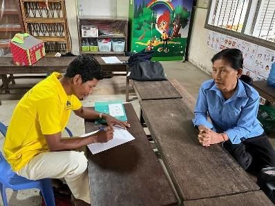 A staff member in yellow and a teacher sit at a wooden desk filling in a survey form