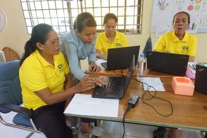 Teachers in yellow shirts practise on laptops at a table while a trainer in blue guides them