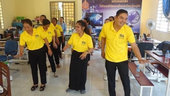 Teachers and trainers in yellow shirts stand in a line and move together in a workshop room
