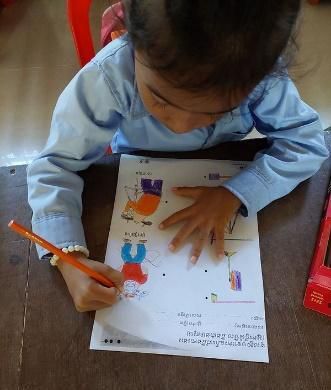A young child colours in a printed worksheet with an orange pencil at a wooden desk