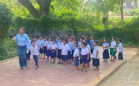 A teacher in blue leads a line of young pupils in school uniform in an outdoor activity