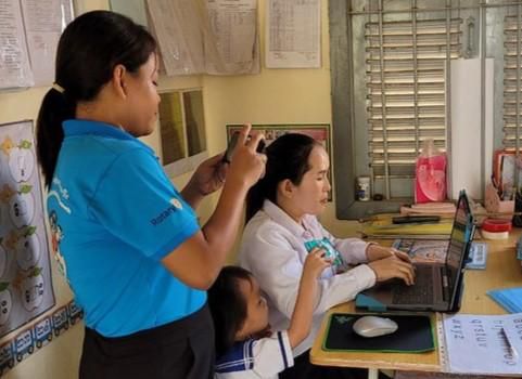 A project staff member helps a teacher type on a laptop during the computer skills check