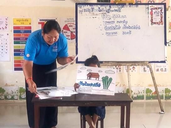 A teacher holds up a picture card in front of a whiteboard to teach a preschool lesson