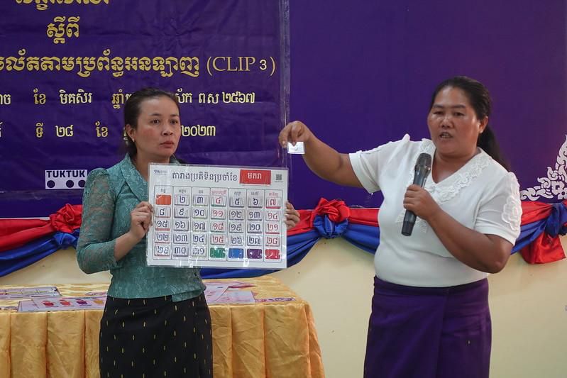 A trainer holds up a classroom decoration chart while a colleague speaks at the pre-project workshop