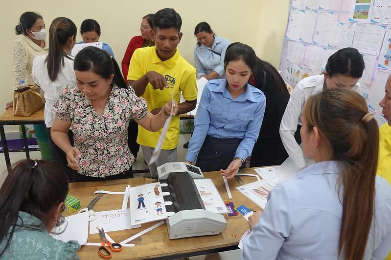 Teachers gather around a laminating machine to finish decorative teaching tools at the pre-project workshop