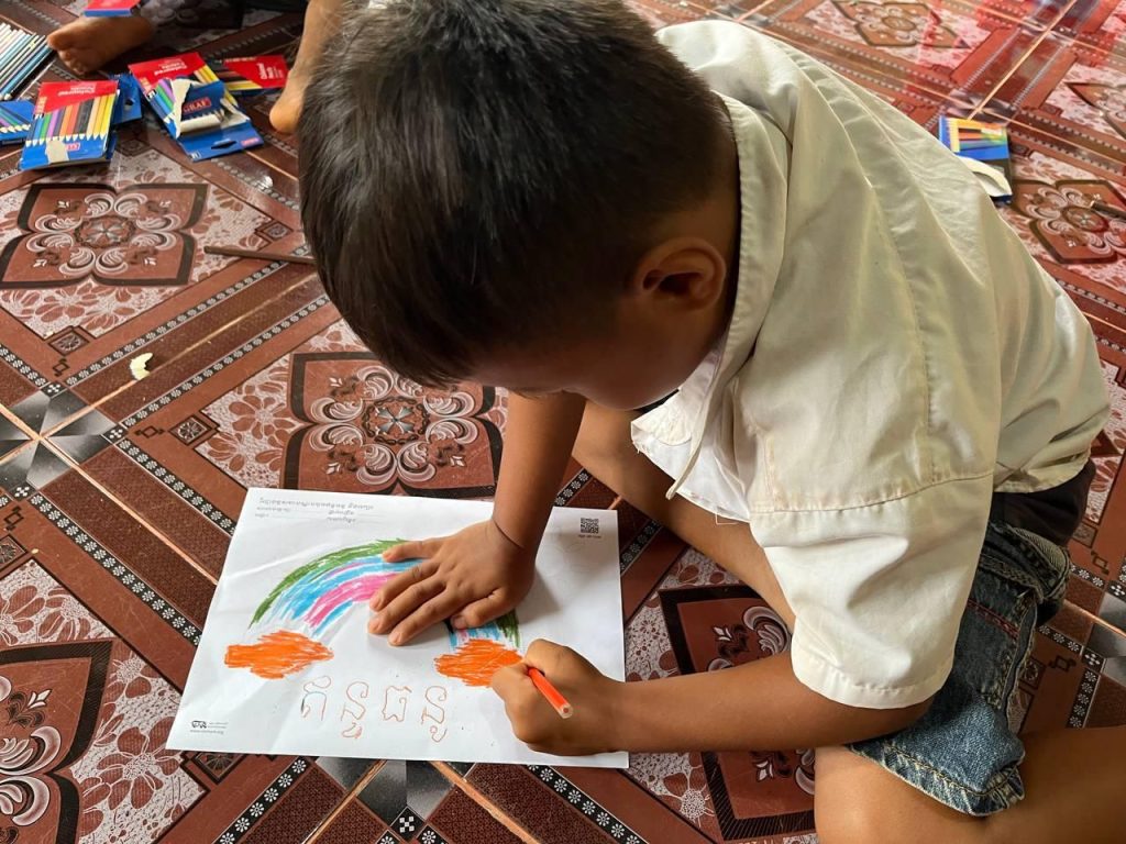 A preschool child kneels on a patterned mat coloring a rainbow worksheet with an orange crayon
