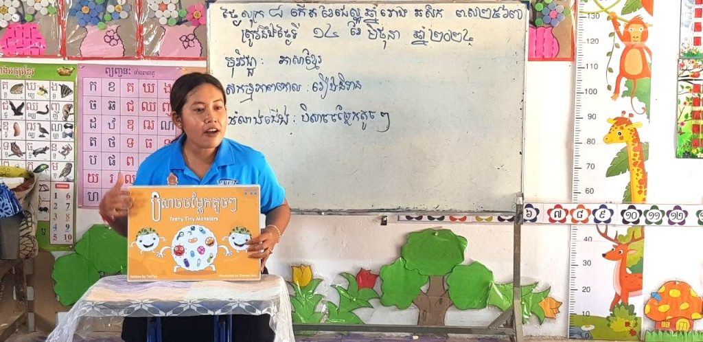 A teacher in a blue shirt shows a Kamishibai picture card to the class during a lesson