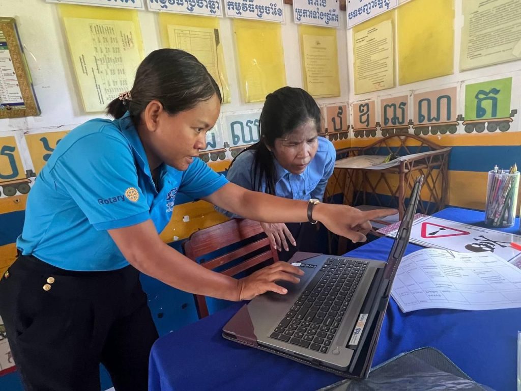 A woman in a blue shirt works on a laptop at a classroom table while another person watches