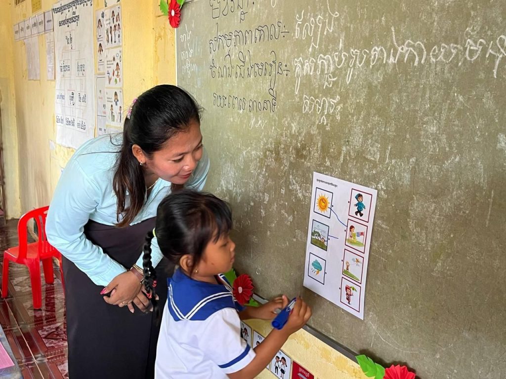 A teacher helps a young girl match picture cards pinned to the classroom wall