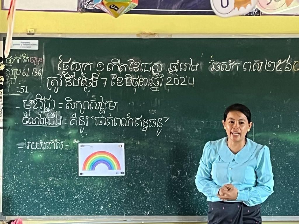 A teacher stands before a chalkboard with a rainbow picture taped beside the day’s lesson notes