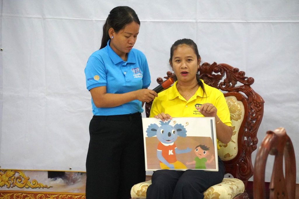 A seated teacher in yellow shows an illustrated Kamishibai card while a colleague holds the microphone