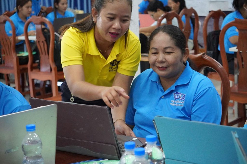 A woman in a yellow polo points at a laptop screen while a seated teacher follows along