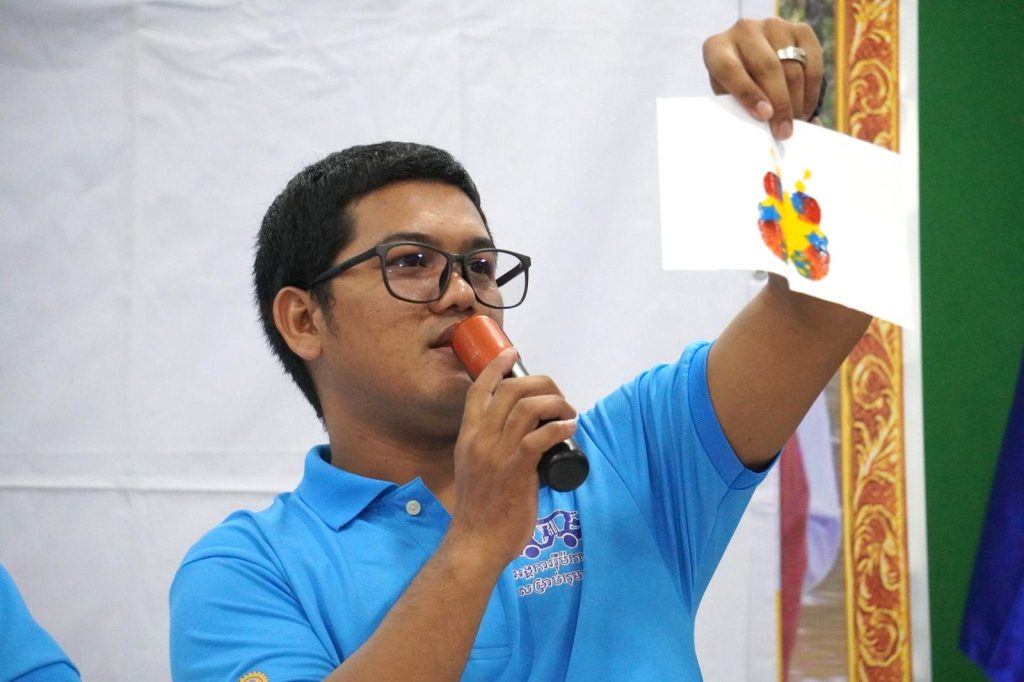 A man in glasses speaks into a microphone while holding up a colorful printed card