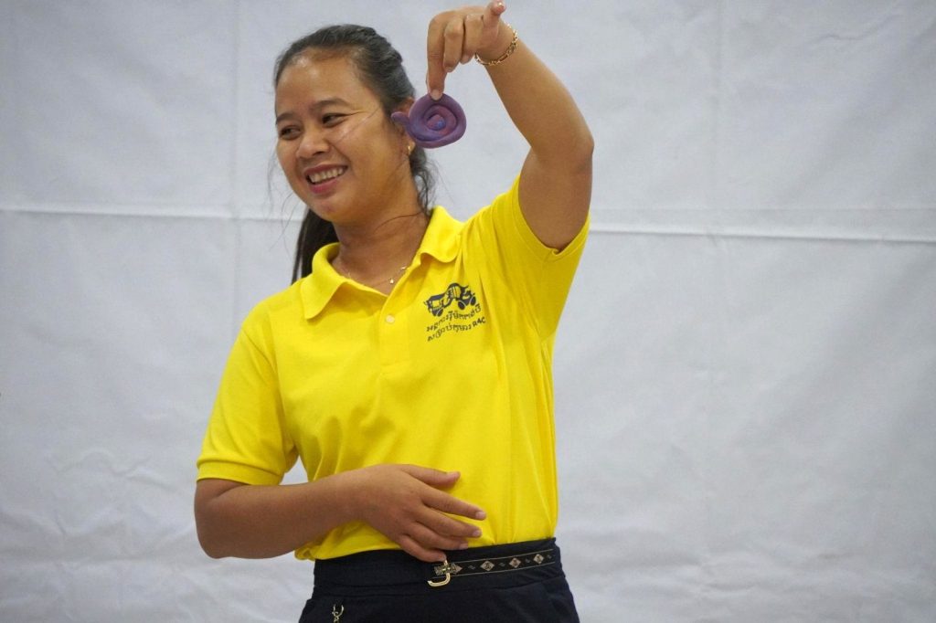 A teacher in a yellow polo smiles and holds up a purple object during the workshop demonstration