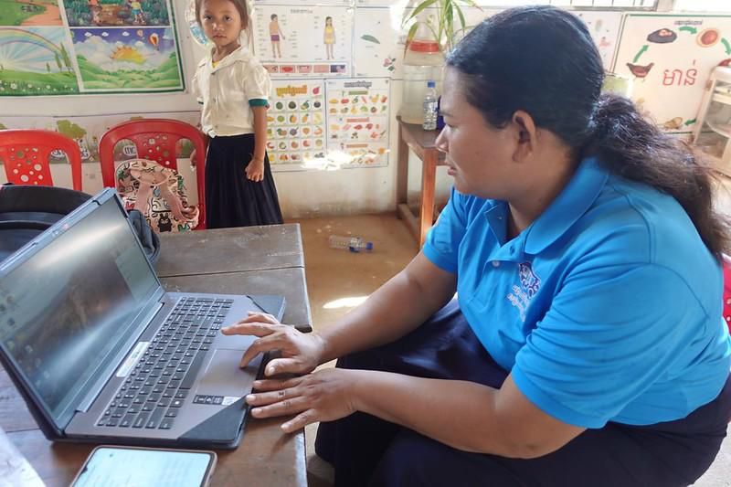 A teacher in a blue shirt types on a laptop at a desk in a decorated classroom