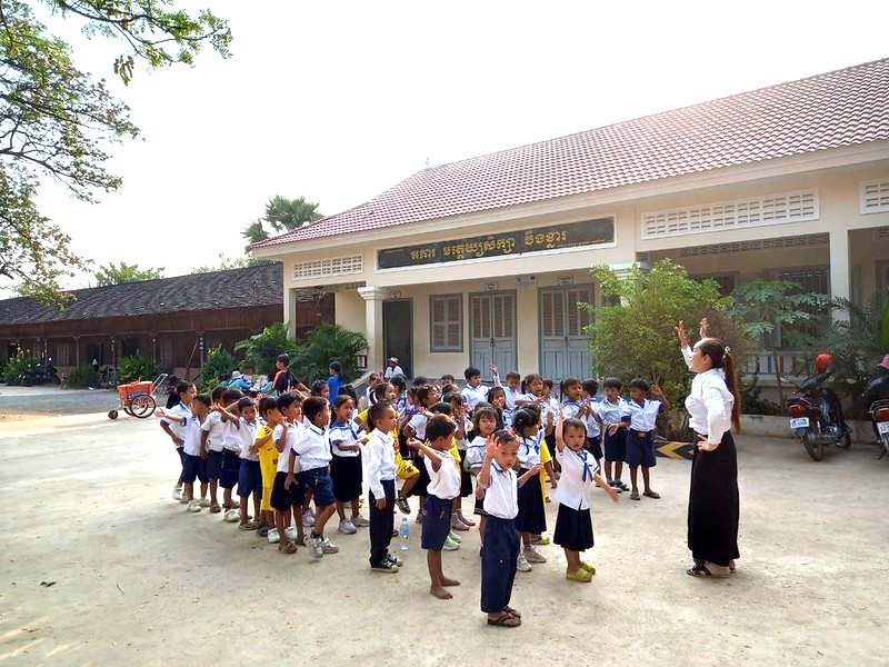 A teacher leads a group of children in school uniforms in a song and movement activity in a schoolyard