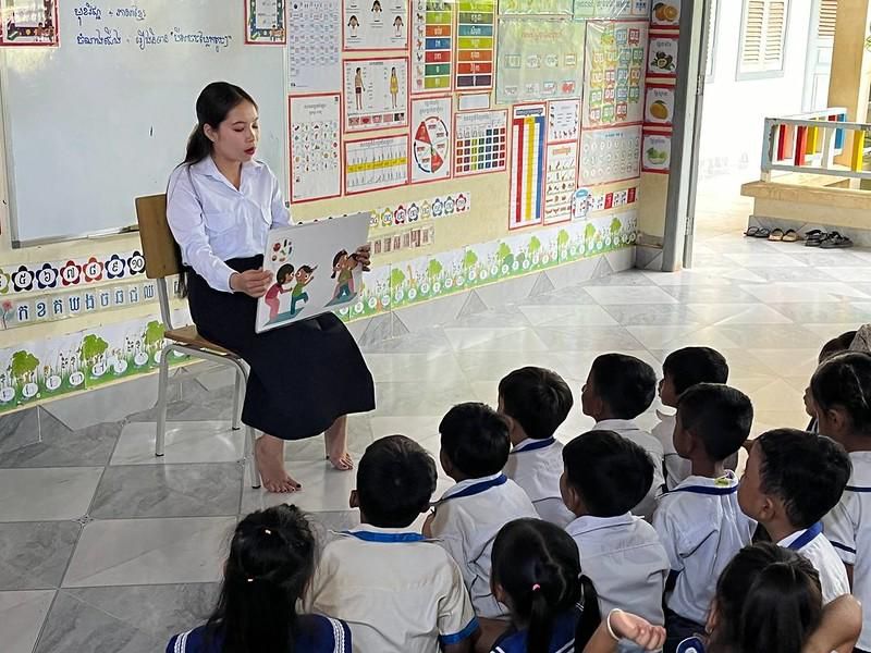 A teacher sits in front of preschool children and reads a storybook aloud in a bright classroom