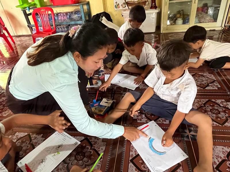 A teacher bends over to help seated children with worksheets on a patterned floor mat