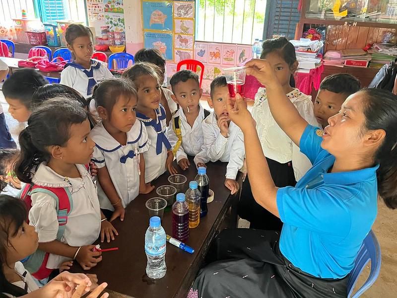 Children crowd around a teacher who holds up a tube while bottles of colored water sit on the table
