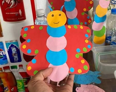 A hand holds a red paper butterfly craft with a caterpillar body decorated with colored dots