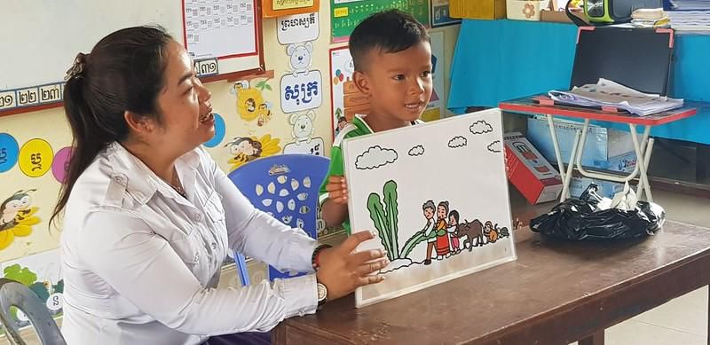A teacher in white shows a Kamishibai picture card to a boy seated at a classroom table