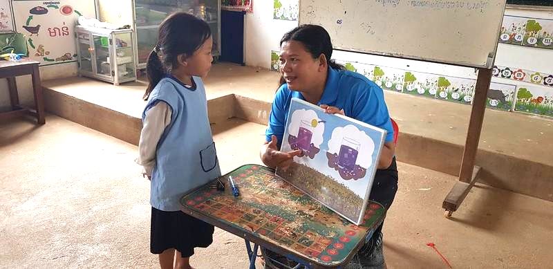 A teacher shows a Kamishibai card to a girl standing beside a classroom table