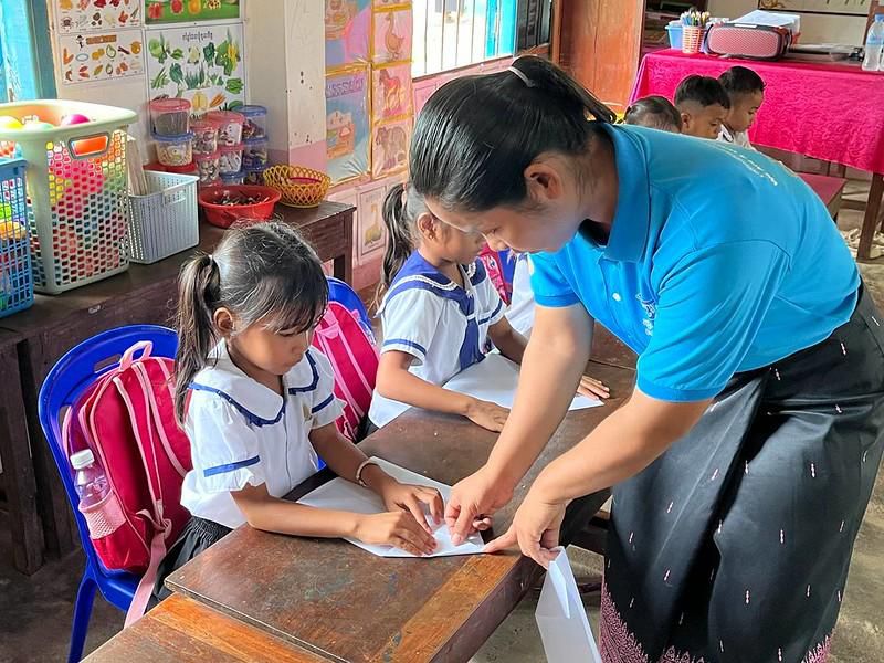 A teacher helps a girl at her desk fold a sheet of paper into a craft
