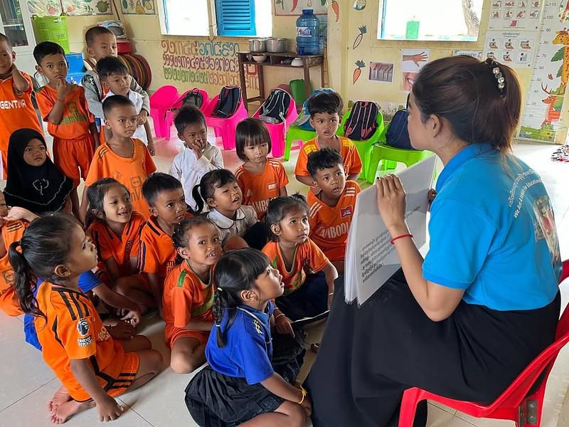 A teacher reads aloud to children in orange uniforms sitting on the floor of a classroom