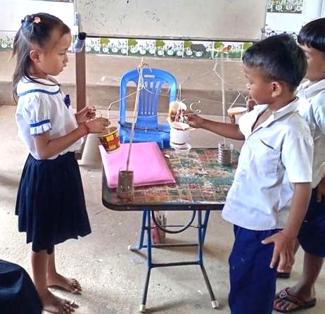 Children in school uniforms gather around a table to watch a simple science experiment with cups