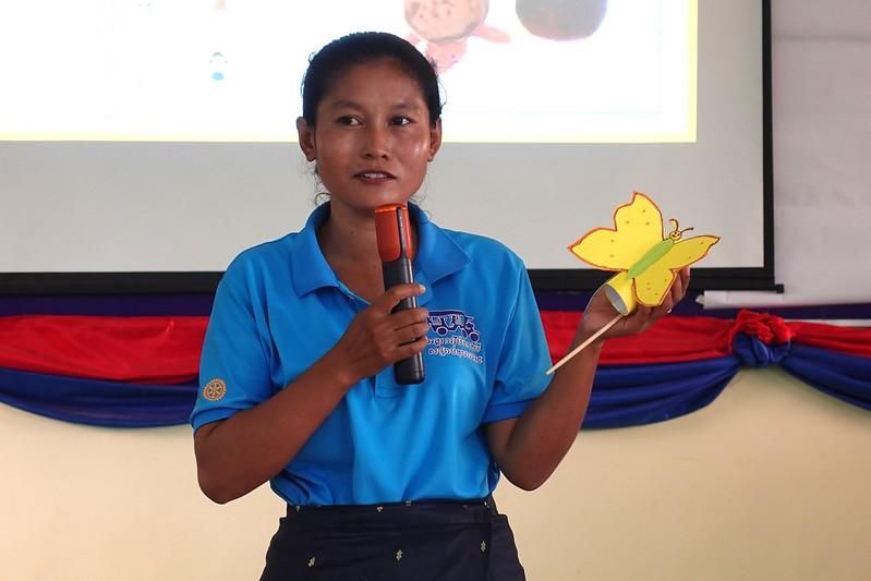 A teacher speaks into a microphone and holds up a yellow paper butterfly craft