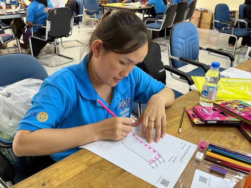 A teacher in a blue shirt draws a craft pattern in pencil at a table with colored pencils