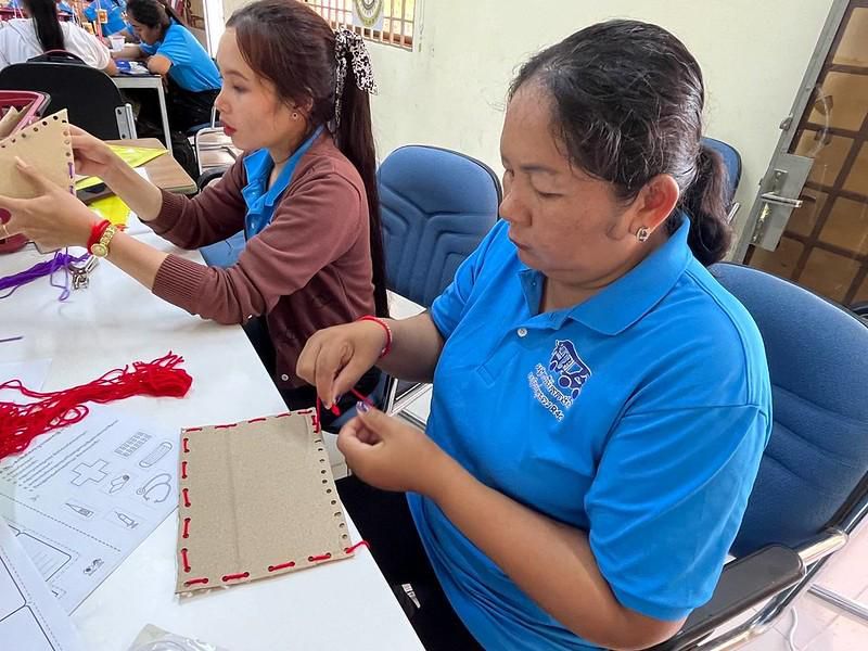 Two teachers sew and thread a cardboard craft at a table covered with patterns and red yarn