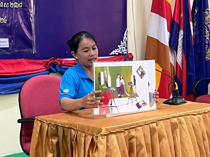 A teacher in a blue shirt holds up a Kamishibai card with a colorful illustration at a table