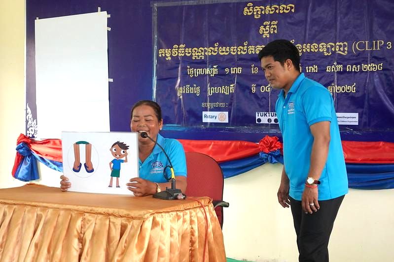 A teacher reads a Kamishibai story card while a man in a blue shirt stands beside the stage