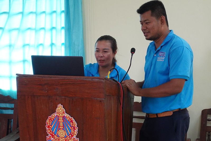 A woman and a man in blue shirts stand at a lectern while she speaks into the microphone