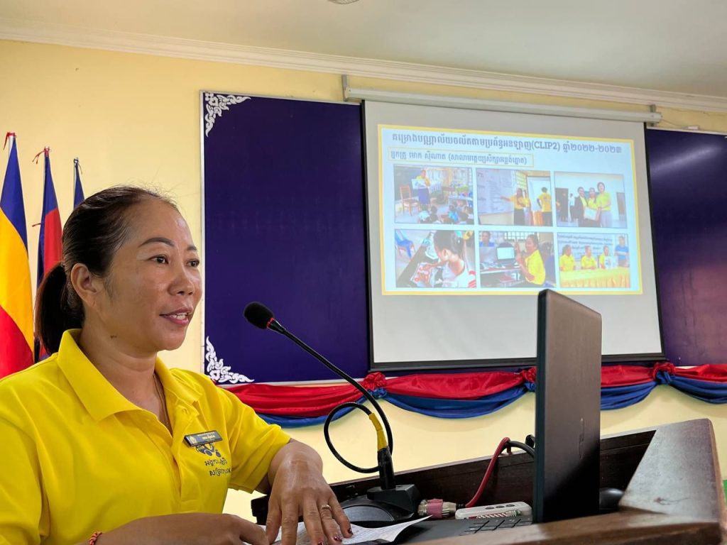 A woman in a yellow polo smiles beside a microphone as a presentation of photos shows on screen