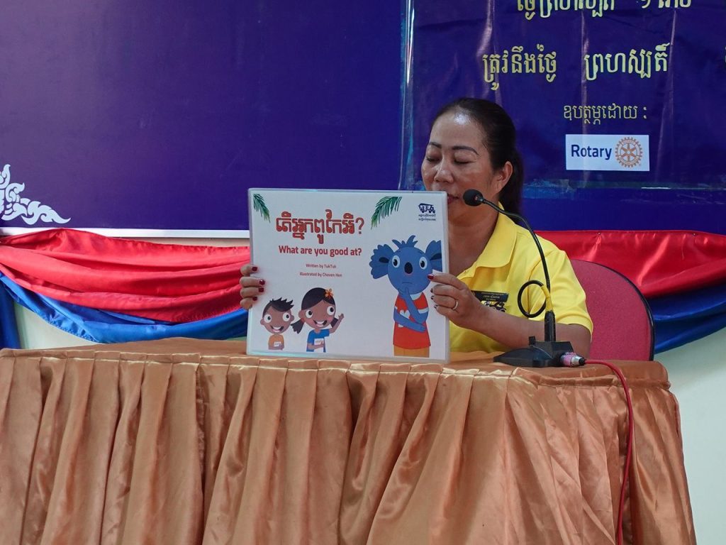 A teacher in a yellow polo holds up the storybook What Are You Good At? at a table