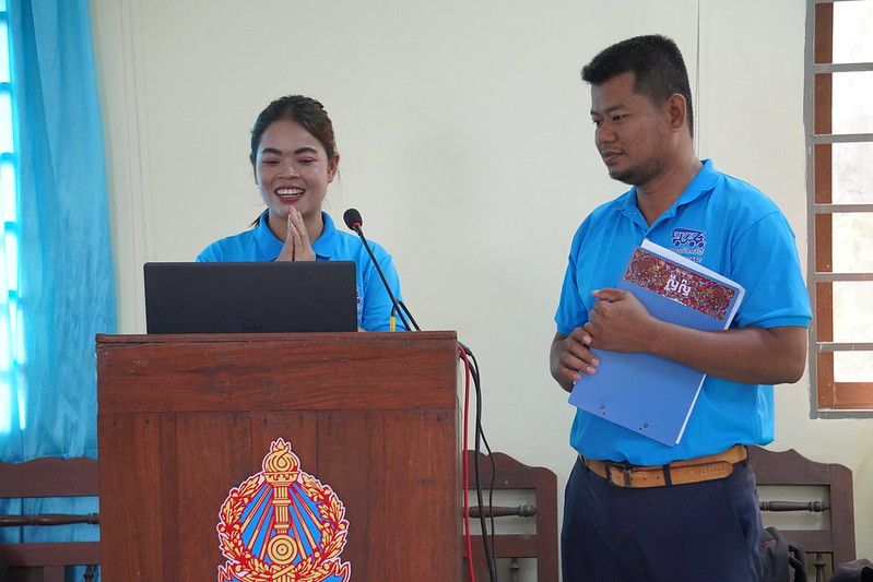 A smiling woman in a blue shirt stands at a lectern with a man holding a folder beside her