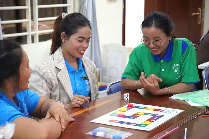 Teachers laugh together while playing a board game with dice at a wooden table