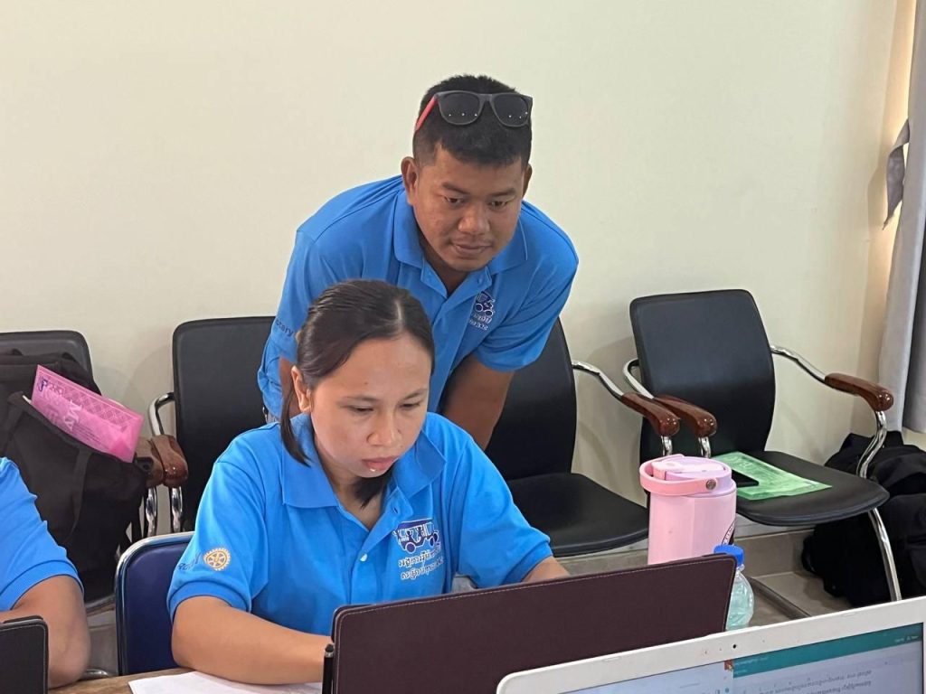 A teacher at a laptop receives help from a man in a blue shirt standing behind her