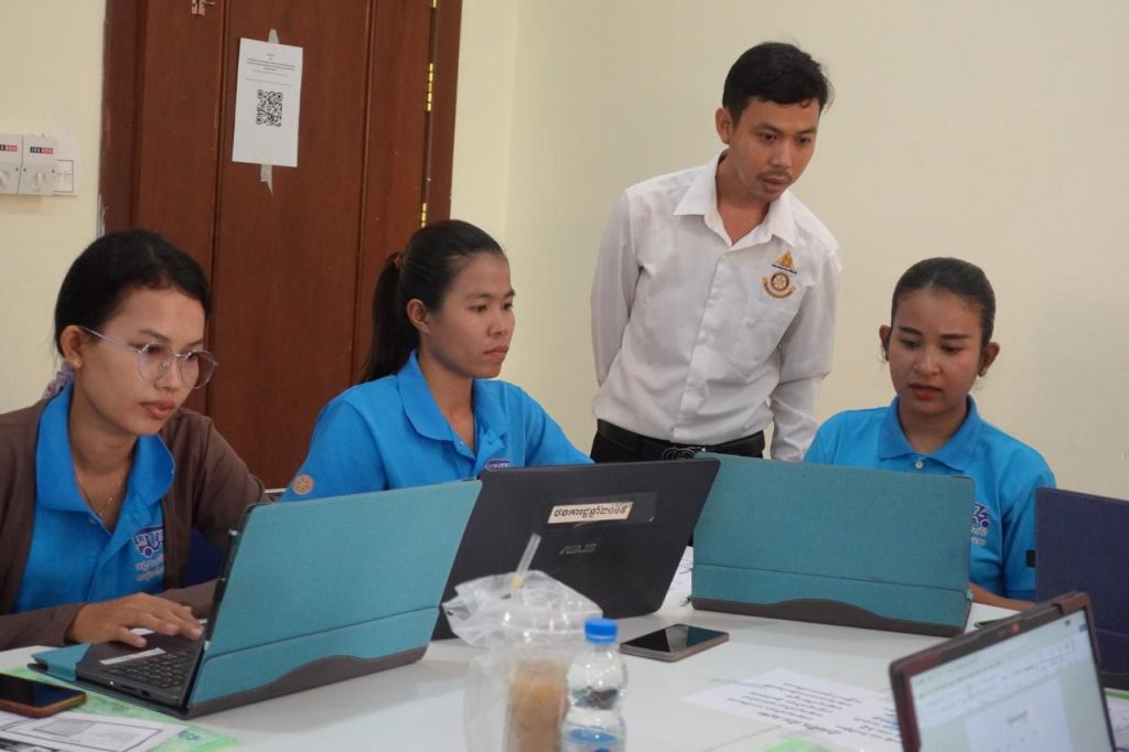 Three teachers in blue shirts work on laptops while a man in a white shirt looks on