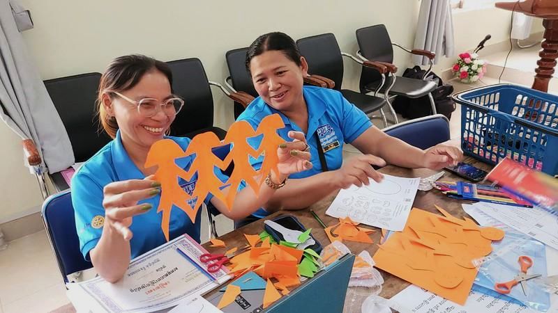 Two teachers smile as they hold up a chain of orange paper figures cut for the craft program