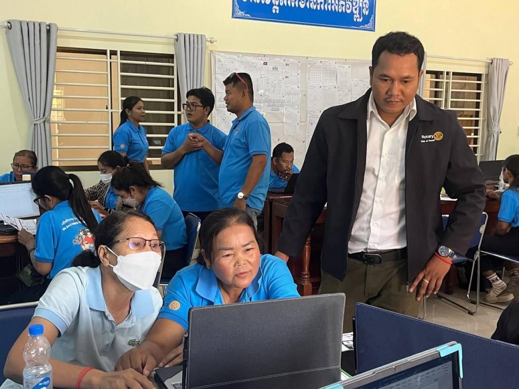 A man in a dark jacket stands beside teachers working at laptops in a crowded training room