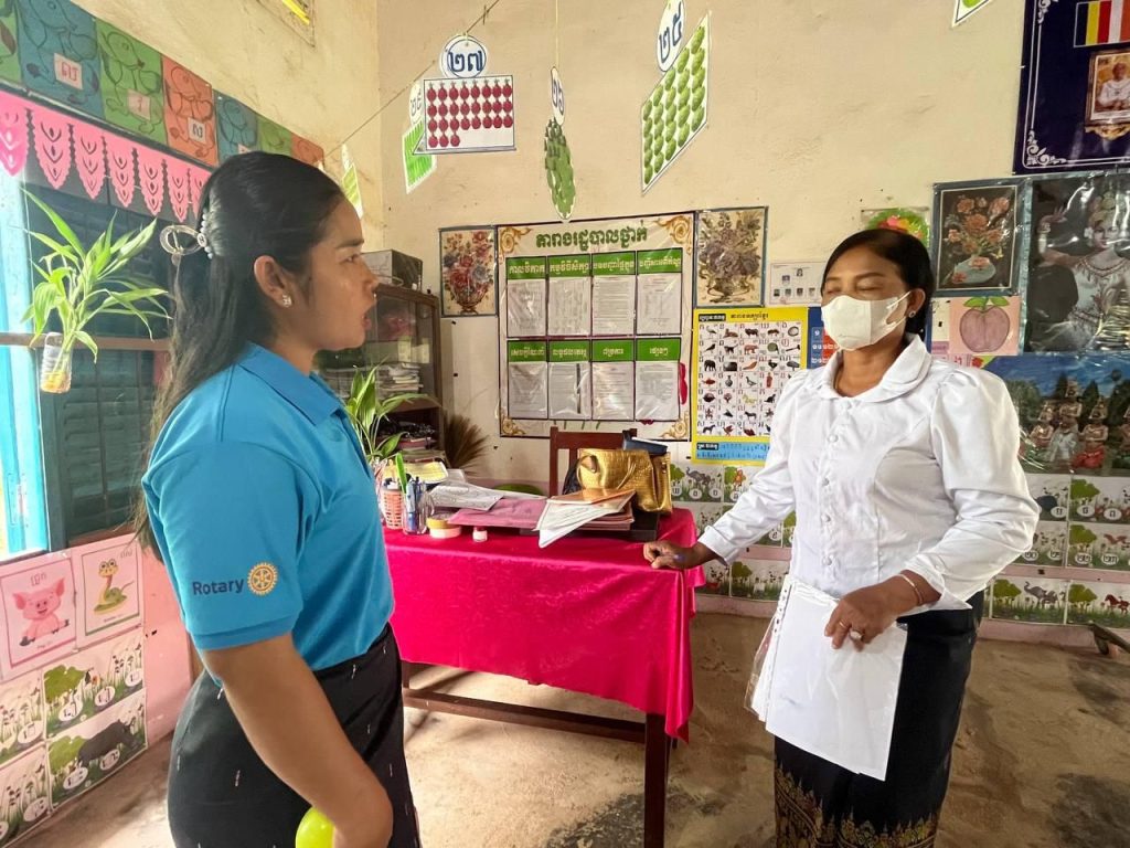 A teacher and an assessor in a face mask stand beside a classroom display table during an assessment