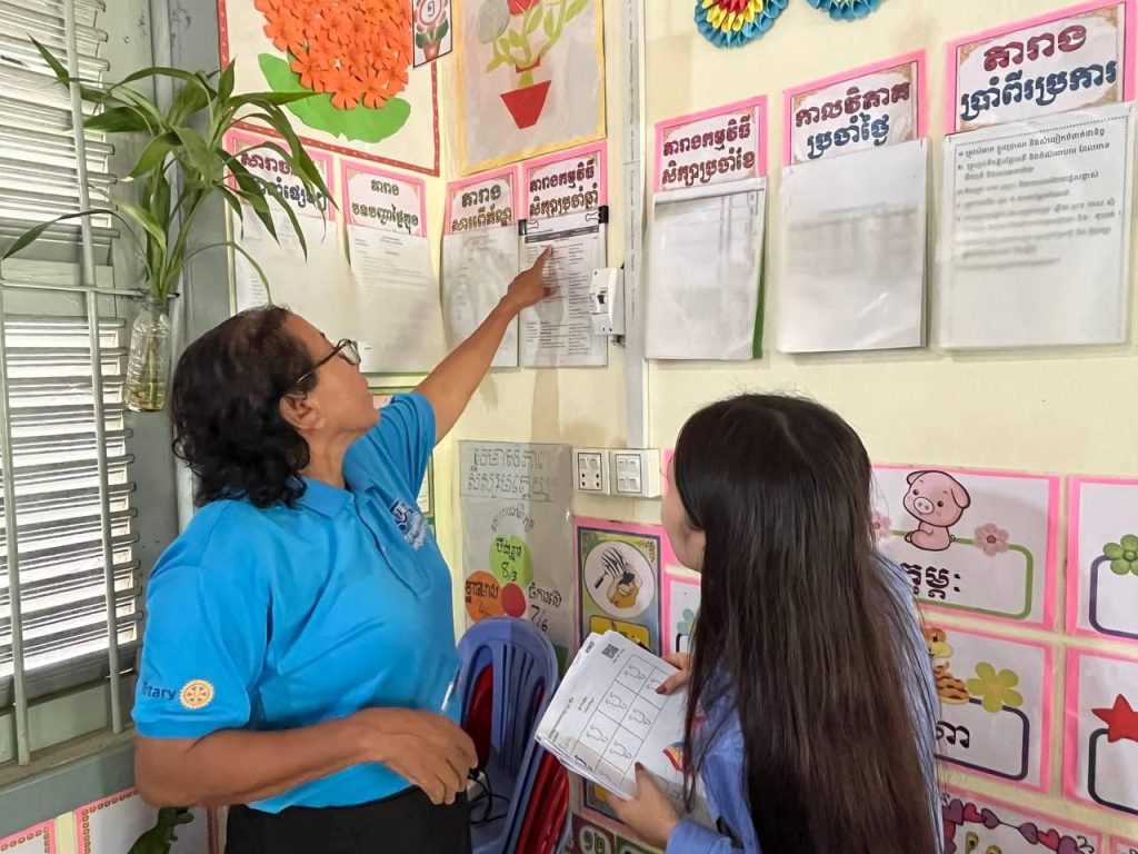 A teacher points to wall charts in a preschool classroom while an assessor holds a form