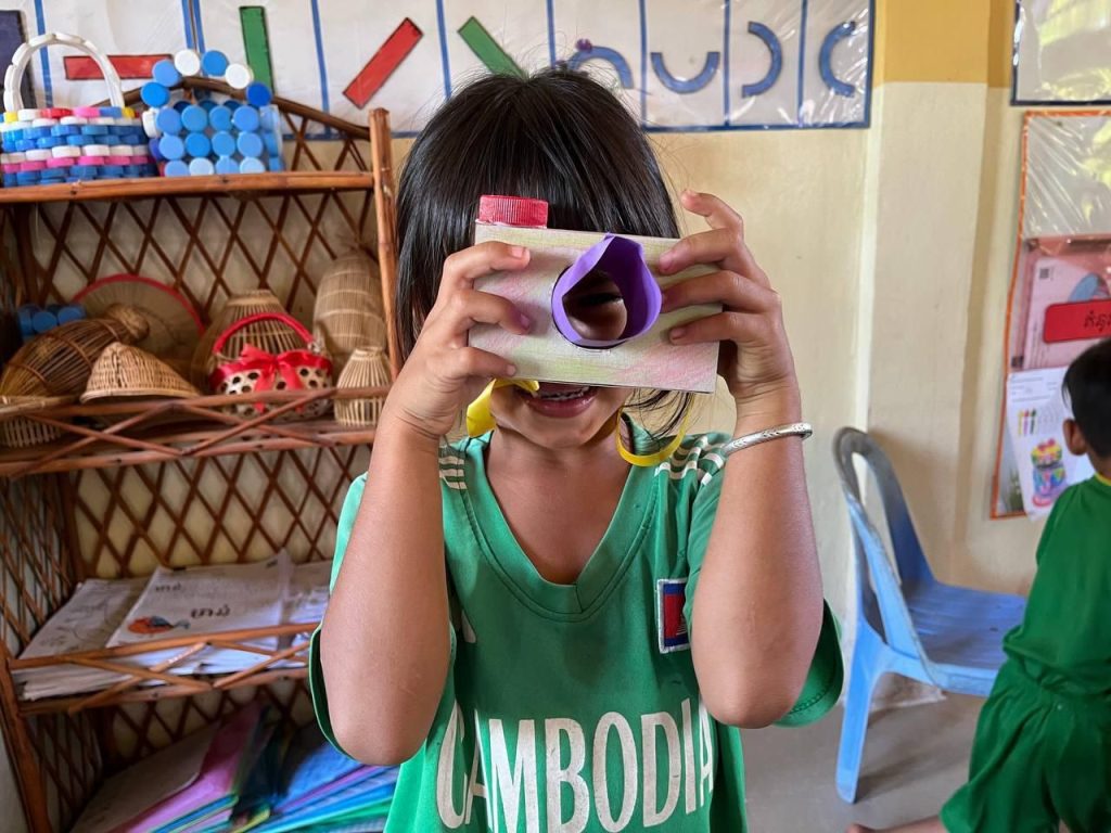 A young child in a green Cambodia shirt holds a paper camera craft in front of their face