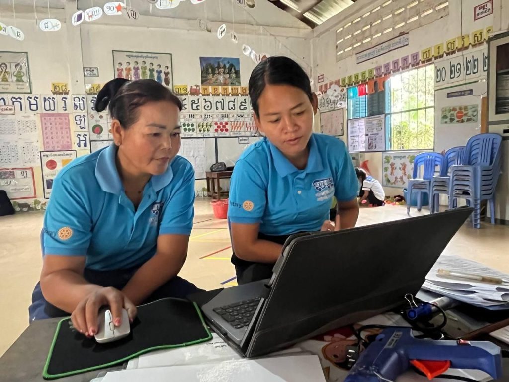 Two teachers use a laptop together on the classroom floor during a follow-up school visit